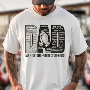 May include: White t-shirt with a black graphic design featuring the word "DAD" and religious text. The design includes praying hands and various Bible verses. The shirt's message is "MAN OF GOD-PROTECTOR-HERO."