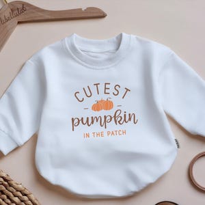 May include: White baby bodysuit with the text "Cutest Pumpkin in the Patch" in brown and orange. The bodysuit is displayed on a wooden hanger. A small embroidery hoop and a woven basket are in the background.