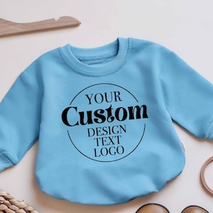 May include: A light blue baby sweatshirt with the text "YOUR Custom DESIGN TEXT LOGO" in a black circular design. The sweatshirt is displayed on a light background with a wooden hanger.
