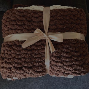 May include: A folded brown chunky knit blanket tied with a beige ribbon. The blanket has a textured, woven appearance. The ribbon is tied in a bow. The blanket is a cozy home decor item.