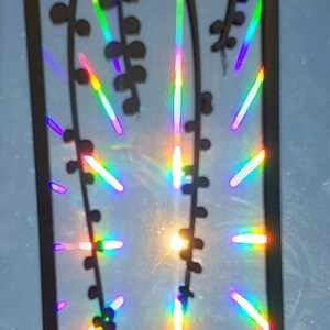 May include: A rectangular wooden cutout with a handle, featuring a floral design. The cutout is held up to the sun, creating a rainbow effect with vibrant colors. The design includes a series of small, circular shapes.