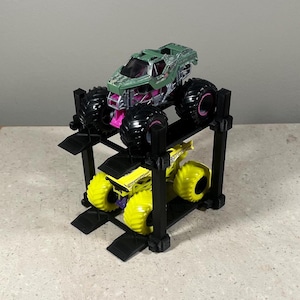 May include: Two toy monster trucks on a black plastic stand. The top truck is green and black with pink accents. The bottom truck is yellow and black with purple accents.