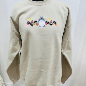 Op de afbeelding: Een beige sweatshirt met een geborduurd beeld van Totoro en de roetgeesten uit de film Mijn buurman Totoro.