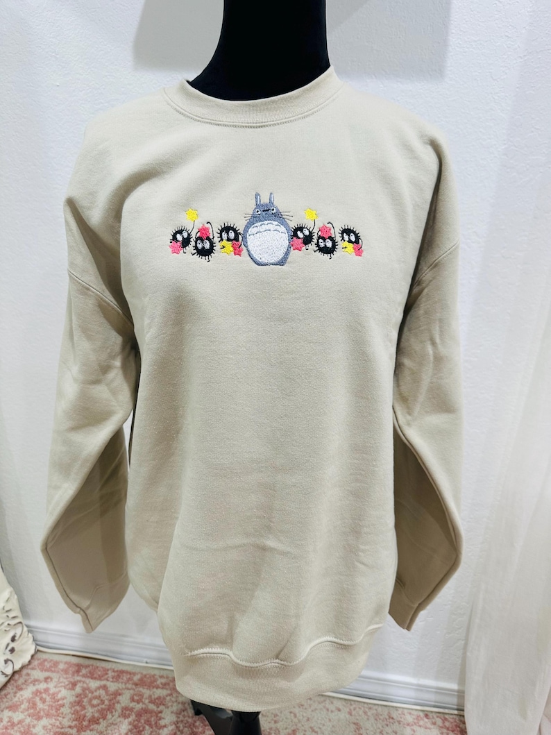 Op de afbeelding: Een beige sweatshirt met een geborduurd beeld van Totoro en de roetgeesten uit de film Mijn buurman Totoro.