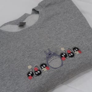 Op de afbeelding: Grijs sweatshirt met een geborduurd beeld van Totoro en de roetgeesten uit de film Mijn buurman Totoro. De roetgeesten houden sterren vast en staan in een rij met Totoro.