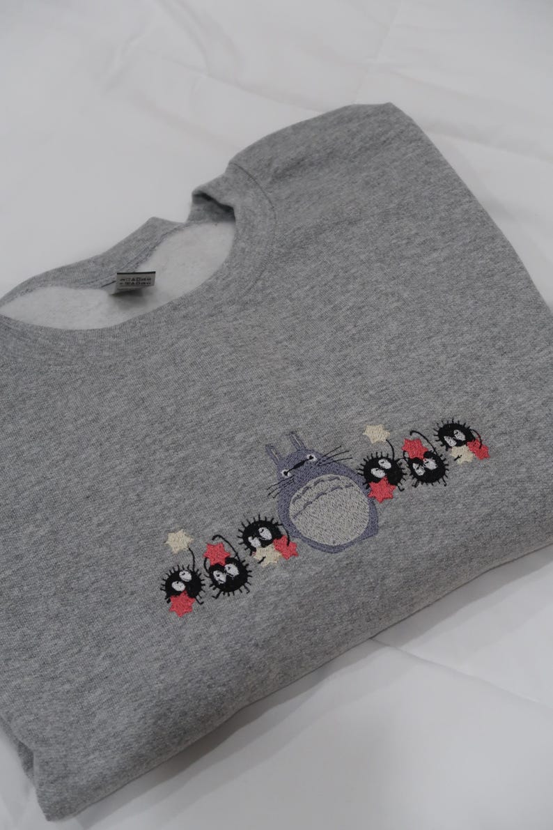 Op de afbeelding: Grijs sweatshirt met een geborduurd beeld van Totoro en de roetgeesten uit de film Mijn buurman Totoro. De roetgeesten houden sterren vast en staan in een rij met Totoro.