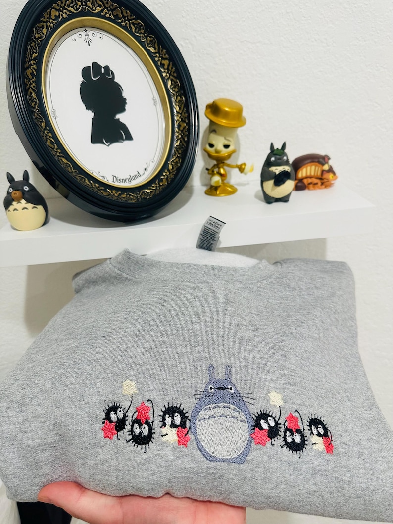 Op de afbeelding: Grijs sweatshirt met een geborduurd beeld van Totoro en de roetgeesten uit de film Mijn buurman Totoro. De roetgeesten houden sterren vast en staan voor Totoro.