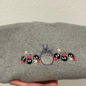 Op de afbeelding: Grijs sweatshirt met een geborduurd beeld van Totoro uit de film Mijn buurman Totoro. Totoro wordt omringd door kleine, zwarte, harige wezens met rode accenten.