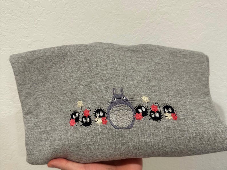 Op de afbeelding: Grijs sweatshirt met een geborduurd beeld van Totoro uit de film Mijn buurman Totoro. Totoro wordt omringd door kleine, zwarte, harige wezens met rode accenten.