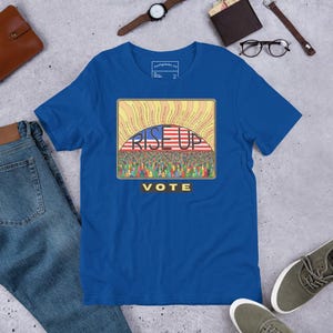 May include: A royal blue t-shirt featuring a graphic print of a crowd of people beneath a rising sun with an American flag pattern. The text RISE UP is printed in black across the center, and the word VOTE is printed in yellow on a black bar.