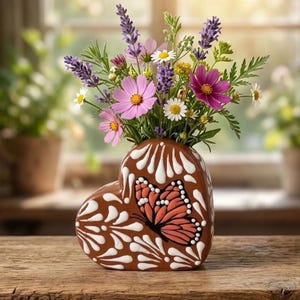 May include: A heart-shaped ceramic vase with a brown base, decorated with white painted swirls and a prominent orange and black butterfly design. The vase is filled with a colorful arrangement of purple lavender, pink cosmos, and white daisies.