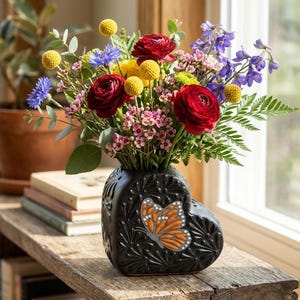 May include: A black heart-shaped vase with a textured surface and a painted orange and black butterfly, filled with a colorful bouquet of red ranunculus, yellow billy balls, blue cornflowers, purple delphinium, and small pink flowers.