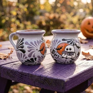 May include: Two round ceramic mugs with a white base, featuring a hand-painted design of a ghost holding a mug in front of an orange moon, surrounded by black bats and purple floral patterns. The mugs have a wide rim and a glossy finish.