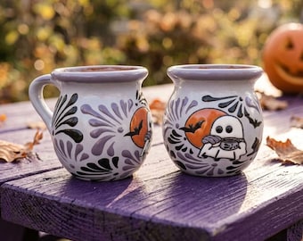 Puede incluir: Dos tazas de cerámica redondas con base blanca, con un diseño pintado a mano de un fantasma sosteniendo una taza frente a una luna naranja, rodeado de murciélagos negros y patrones florales morados. Las tazas tienen un borde ancho.