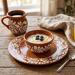 May include: A terracotta-colored ceramic mug, bowl, and plate set featuring a white hand-painted floral pattern. The bowl contains yogurt topped with fresh blueberries and honey, accompanied by a small silver-toned metal spoon.
