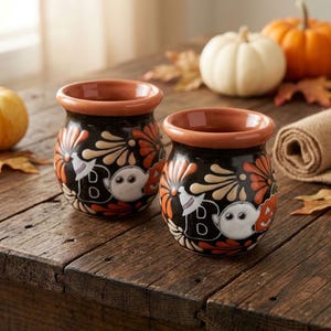 May include: Two round ceramic mugs with a black base, featuring a repeating pattern of white ghosts, orange and cream-colored floral motifs, and the letter B in a bold font. Each mug has a wide, smooth, terracotta-colored rim and opening.