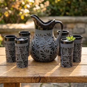 May include: A ceramic pitcher and five matching tumblers featuring a matte gray finish with a glossy black rim and a repeating black embossed fan-like pattern. The set is arranged on a wooden surface, with one tumbler containing ice and mint.