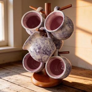 May include: A wooden mug tree stand holding four ceramic mugs and one matching creamer pitcher. Each piece features a white exterior with a blue bubble-like pattern and a solid light pink interior. The stand has a round base and six wooden arms.