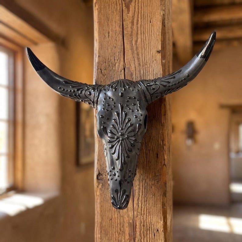 May include: A decorative, dark gray bull skull with intricate floral and dot patterns. The horns are black-tipped. The skull is mounted on a weathered wooden post, creating a rustic, southwestern aesthetic. The skull is a decorative item.