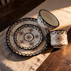 May include: A cream-colored ceramic dinnerware set with a floral design in red and black. The set includes plates, a bowl, and a mug. The plates have a diameter of approximately 10 inches.