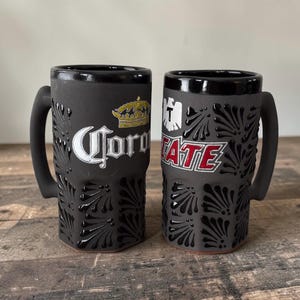 May include: Two black ceramic beer mugs with handles. One mug features the word "Corona" with a gold crown, the other has the word "ATE" in red. Both mugs have a textured, fan-like pattern and are on a wooden surface.