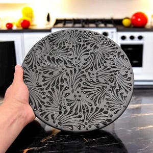 May include: A round, gray decorative plate with a raised, black floral pattern. The plate is held in a hand, showcasing the intricate design. The kitchen background includes a stove and countertop.