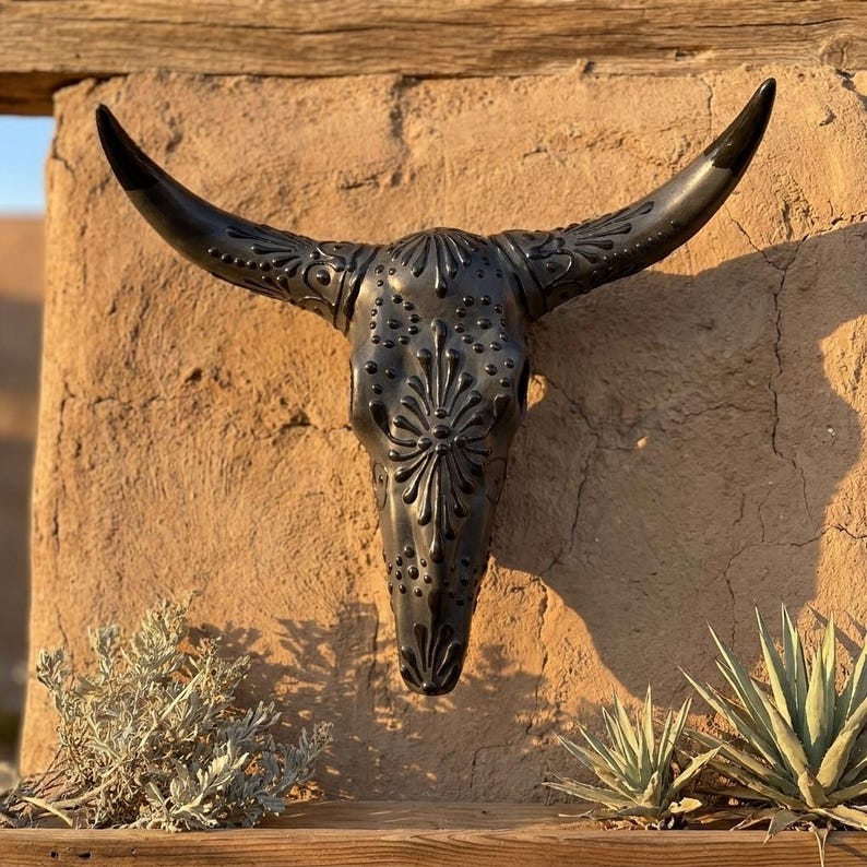 May include: Black decorative steer skull with intricate floral and dot patterns. The skull is mounted on a textured, light brown wall, with the horns curving upwards. The skull is a decorative piece.