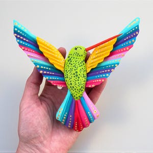 May include: A vibrantly painted wooden hummingbird sculpture with outstretched wings. The bird features a green body with black spots, yellow and rainbow wings, and a red beak. The wings are decorated with gold dots.
