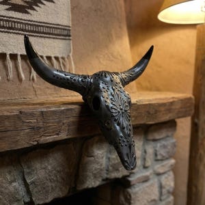 May include: A decorative, dark gray bull skull with intricate floral and dot patterns. The skull has long, curved horns with black tips. It is displayed on a rustic wooden shelf above a stone fireplace, showcasing a southwestern theme.