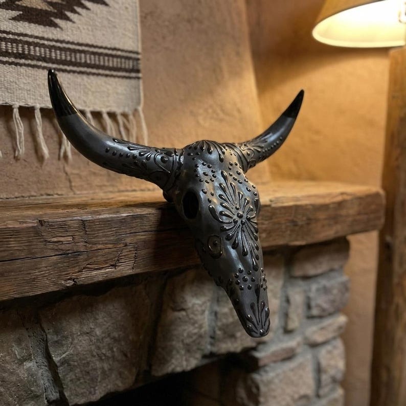 May include: A decorative, dark gray bull skull with intricate floral and dot patterns. The skull has long, curved horns with black tips. It is displayed on a rustic wooden shelf above a stone fireplace, showcasing a southwestern theme.