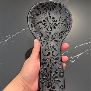 May include: A black ceramic spoon rest with an ornate floral design. The spoon rest is held in a hand and features a raised, glossy pattern. The design includes stylized flowers and leaves, adding a decorative touch to the kitchen utensil.