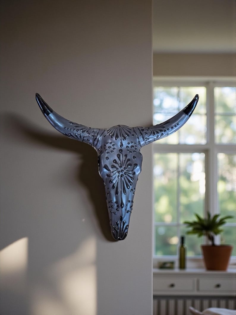 A decorative black Talavera  longhorn skull wall hanging. The skull features intricate embossed detailing and black-tipped horns. The wall art is mounted on a neutral-colored wall, adding a rustic touch to the decoration