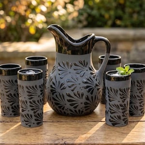 May include: A ceramic pitcher and five matching tumblers in a matte gray finish with a glossy black rim and base. Each piece features a repeating embossed black floral pattern. One tumbler contains ice and a sprig of fresh green mint.