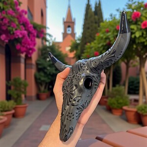 May include: A dark gray decorative bull skull mask with black horns and intricate floral patterns. The mask is held up in front of a blurred background of a building and trees.
