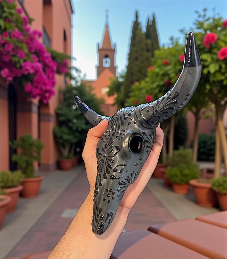 May include: A dark gray decorative bull skull mask with black horns and intricate floral patterns. The mask is held up in front of a blurred background of a building and trees.