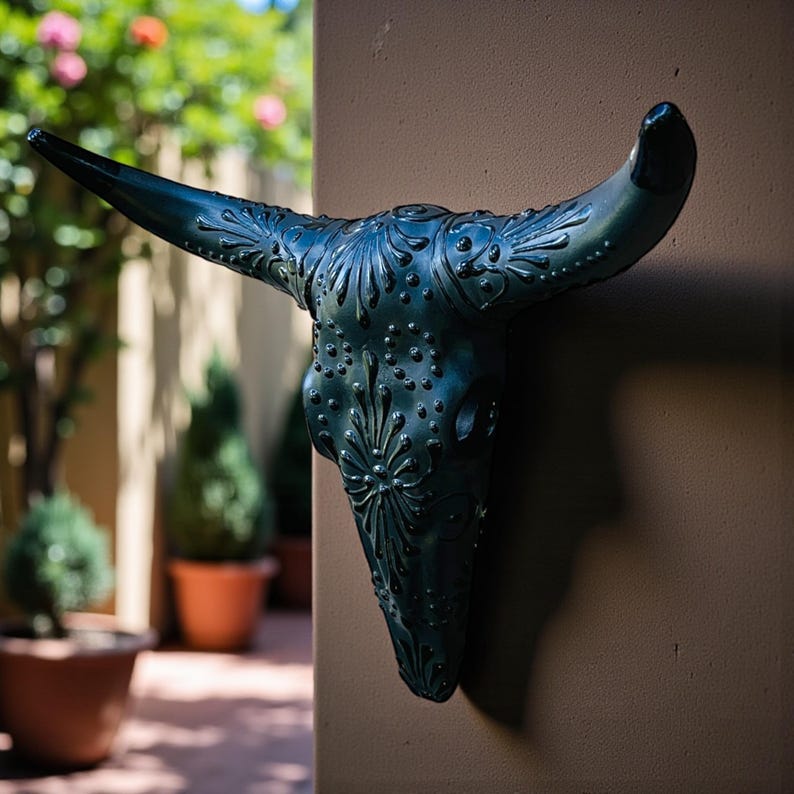 May include: A decorative, dark teal-colored bull skull wall hanging. The skull features intricate embossed floral and dot patterns. The horns are long and curved. The sculpture is mounted on a brown wall, with a blurred outdoor background.