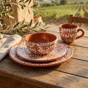 May include: A terracotta dinnerware set consisting of a large plate, a smaller plate, a bowl, and a mug. Each piece features a hand-painted white floral pattern over the reddish-brown clay surface, creating a cohesive look for the table setting.