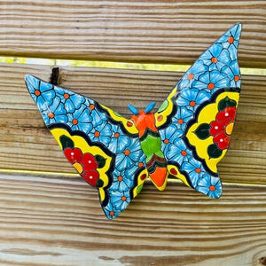 May include: A colorful ceramic butterfly wall hanging. The butterfly features blue wings with orange dots and flower patterns, yellow accents, and red flower details. The body is orange, green, and black. It is mounted on a wooden fence.