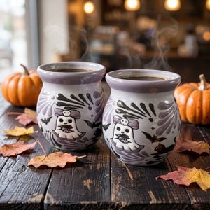 May include: Two round ceramic mugs with a light gray finish, featuring a repeating pattern of white ghosts with purple bows, black bats, and decorative purple swirls. Each mug is filled with a dark liquid and emits a visible wisp of steam.