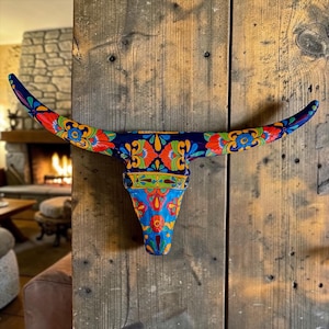 May include: A vibrantly painted bull skull with intricate floral patterns in red, orange, yellow, and blue. The decorative piece is mounted on a rustic wooden wall, showcasing a Southwestern or Mexican folk art style. The horns curve gracefully, adding to its artistic appeal.