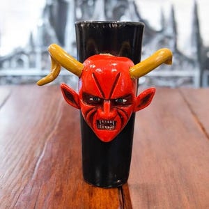May include: A black shot glass with a red devil face and yellow horns. The devil face has blue eyes and a menacing expression. The shot glass is on a wooden surface.