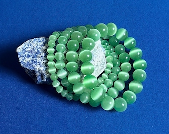 Pode incluir: Um conjunto de quatro pulseiras de pedras preciosas verdes com efeito olho de gato. As pulseiras são feitas de contas redondas e estão empilhadas umas sobre as outras.