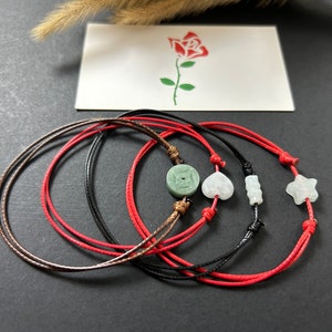 May include: Four red, brown, and black string bracelets with jade and white stone charms. The charms include a circle, a heart, a rectangle, and a star.