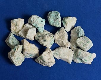 May include: A collection of rough, irregularly shaped stones in shades of white and pale green, scattered on a deep blue surface. The stones have a textured appearance, suggesting a natural mineral composition.