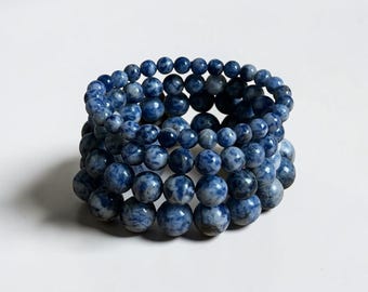 May include: Stack of blue and white beaded bracelets. The bracelets are made of round, polished beads in varying sizes. The beads have a mottled appearance with shades of blue and white. The bracelets are stacked on top of each other.