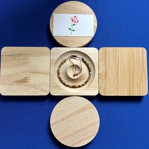 May include: A wooden box with a lid that opens to reveal a small compartment. The compartment contains a brown bracelet with a string closure. The lid of the box has a white card with a red rose illustration.