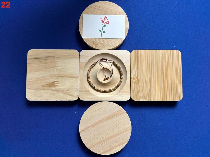 May include: A wooden box with a lid that opens to reveal a small compartment. The compartment contains a brown bracelet with a string closure. The lid of the box has a white card with a red rose illustration.