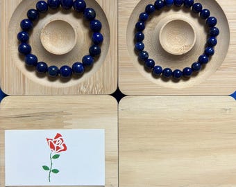May include: Two blue lapis lazuli beaded bracelets on wooden display stands. A small card with a red rose illustration is on a wooden surface.