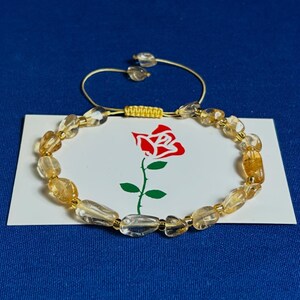 May include: A gold and clear crystal beaded bracelet with a sliding knot closure. The bracelet is on a white card with a red rose illustration.