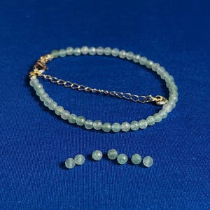 May include: A green gemstone bracelet with a gold clasp. The bracelet is made of small, round beads. There are six extra beads next to the bracelet.